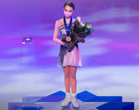 На изображении фигуристка в костюме с медалью на шее стоит с букетом цветов на ледовой арене. Она в белых коньках, а на лице защитная маска. Атмосфера торжественная, сине-фиолетовое освещение создает праздничное настроение